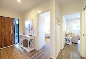 Un interior moderno con una puerta de madera, una consola blanca, una cama y un sofá en una habitación luminosa con piso de madera.