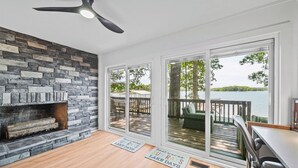 A room with a stone fireplace, a ceiling fan, and large sliding glass doors leading to an outdoor deck with a view of a lake.