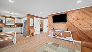 A modern kitchen with white cabinets, a built-in oven, and a refrigerator. There is a dining table with chairs and a TV mounted on the wall.