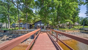 A wooden bridge leading to a building with a deck, surrounded by trees and a body of water.