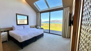 Quarto com cama grande, mesa de cabeceira, cadeira e vista para uma paisagem através de uma porta de vidro deslizante.