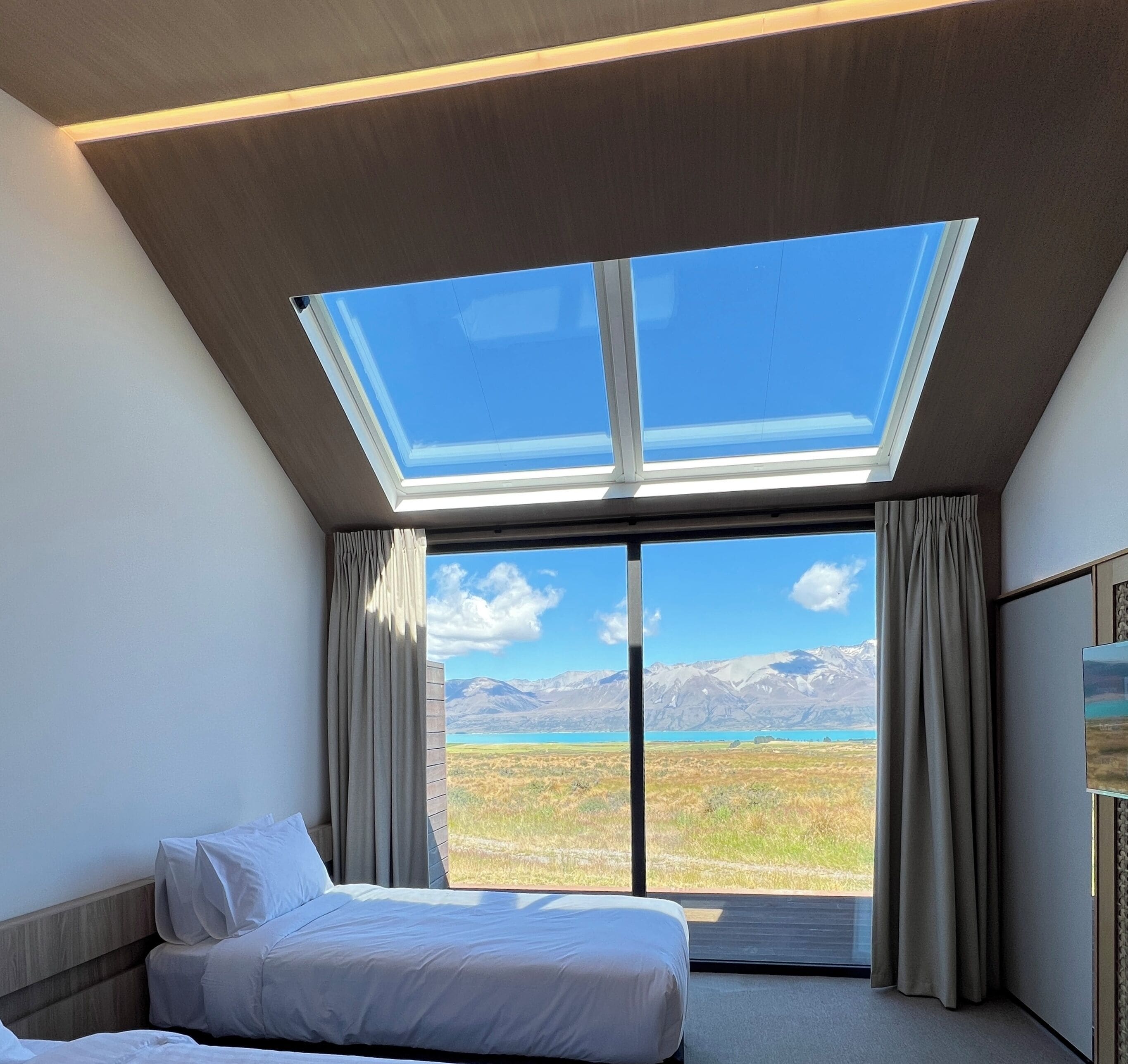 A bedroom with a skylight, a bed, curtains, and a view of mountains and a lake.