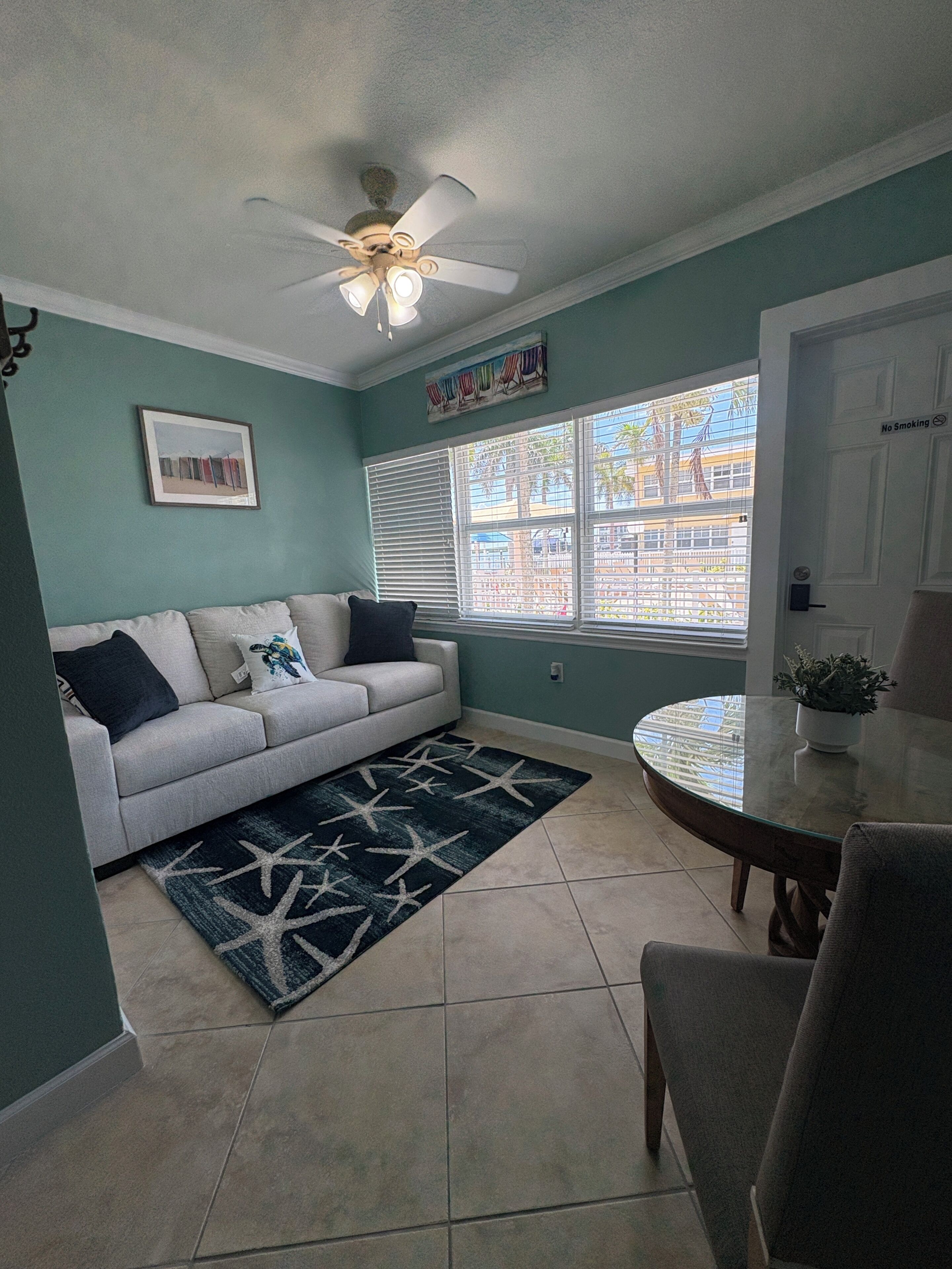 A living room with a sofa, a dining table, a ceiling fan, a window with blinds, and a 'No Smoking' sign.