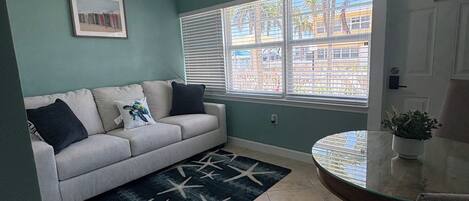 A living room with a sofa, a dining table, a ceiling fan, a window with blinds, and a 'No Smoking' sign.