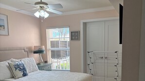 Ein Schlafzimmer mit einem Bett, Deckenventilator, Nachttisch, Lampe, einem Fenster mit Jalousien und einer Tür.