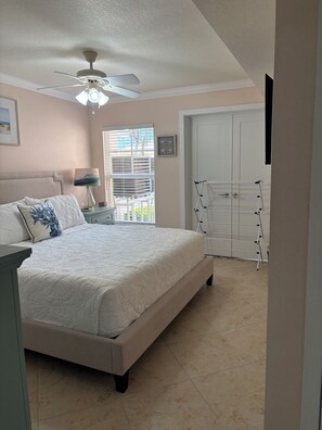 Un dormitorio con cama, ventilador de techo, mesita de noche, lámpara, ventana con persianas y puerta.