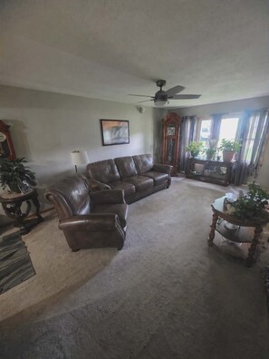 A living room with a brown leather sofa, a ceiling fan, a clock, a painting, and a large window with curtains. - Kansas City World Cup  Home W/ Resort Pool outdoor entertainment area sleeps 8 (Leavenworth)
