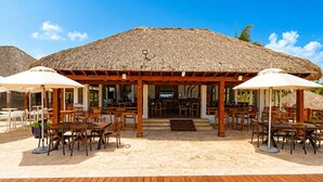 Een restaurant aan het strand met houten tafels en stoelen, een rieten dak en een houten terras.