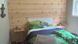 Un dormitorio con techo de madera, una cama con manta verde y azul, una mesita de noche pequeña y una mesita auxiliar.