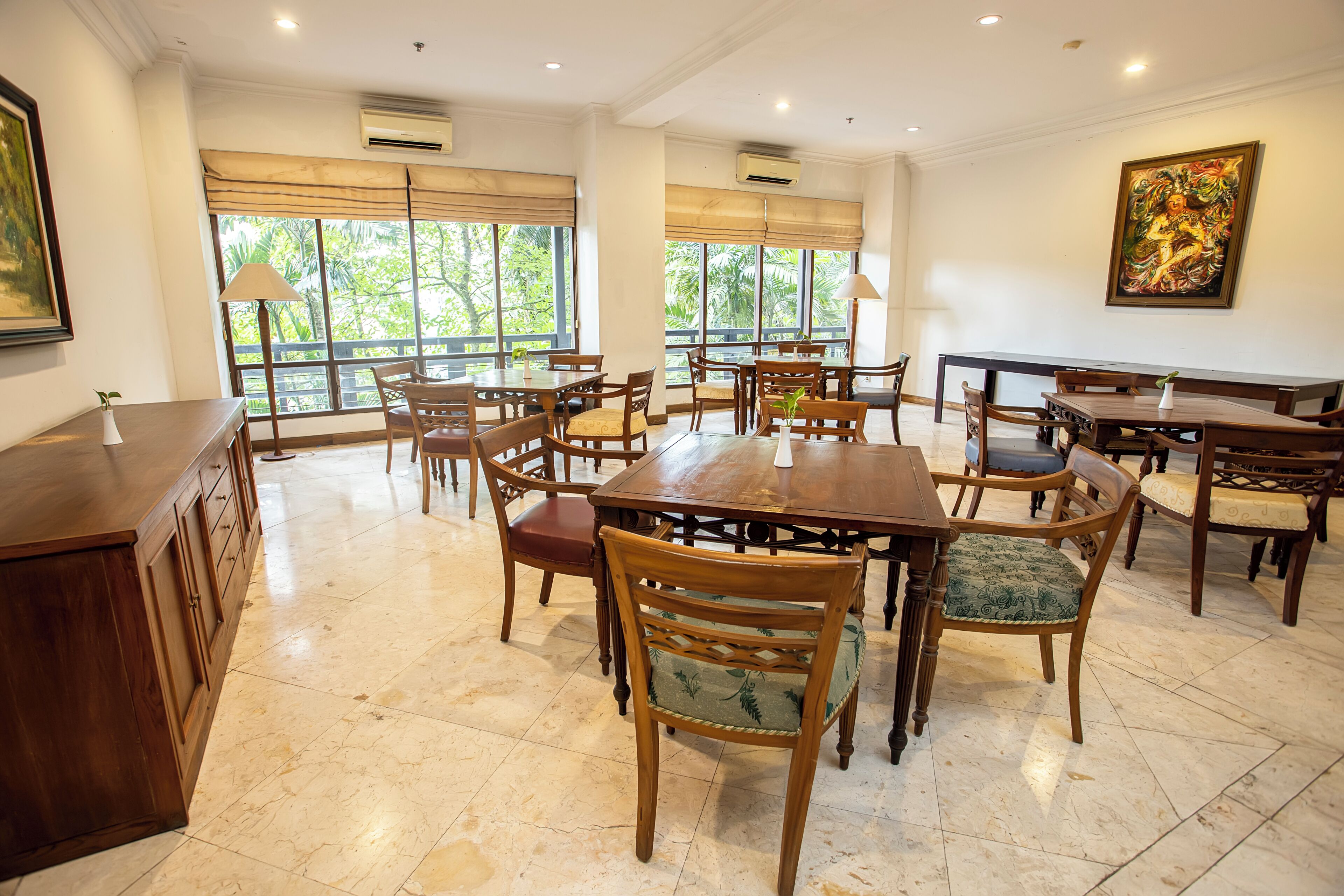 A spacious dining area with multiple wooden tables and chairs, large windows with blinds, and an air conditioning unit on the wall.