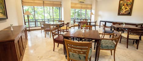 A spacious dining area with multiple wooden tables and chairs, large windows with blinds, and an air conditioning unit on the wall.
