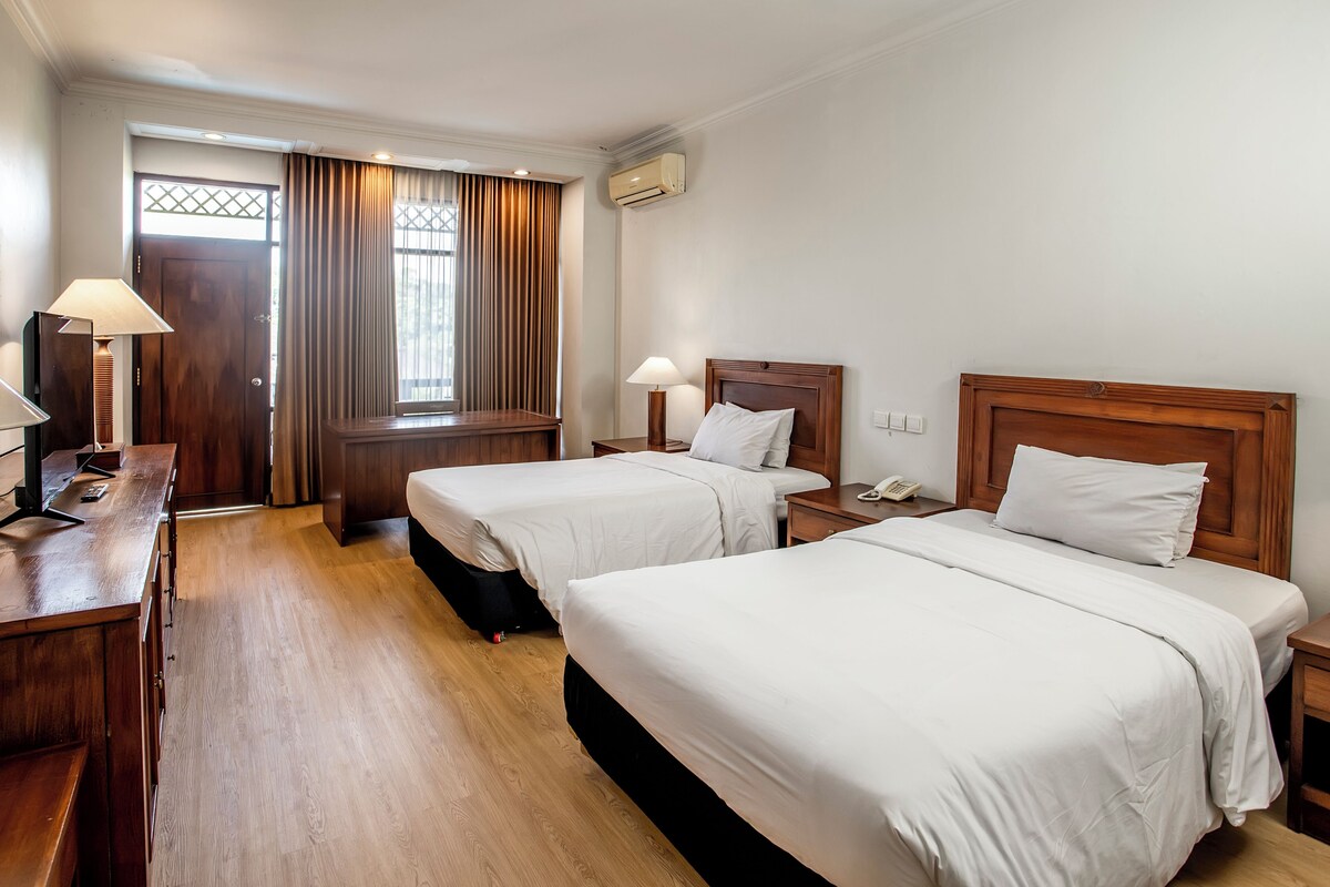 A hotel room with two beds, a desk, a chair, a window with curtains, and an air conditioning unit.