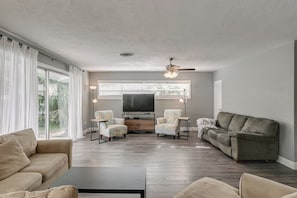 Ein Wohnzimmer mit einer Couch, zwei Sesseln, einem Couchtisch, einem Fernseher und einem Deckenventilator.