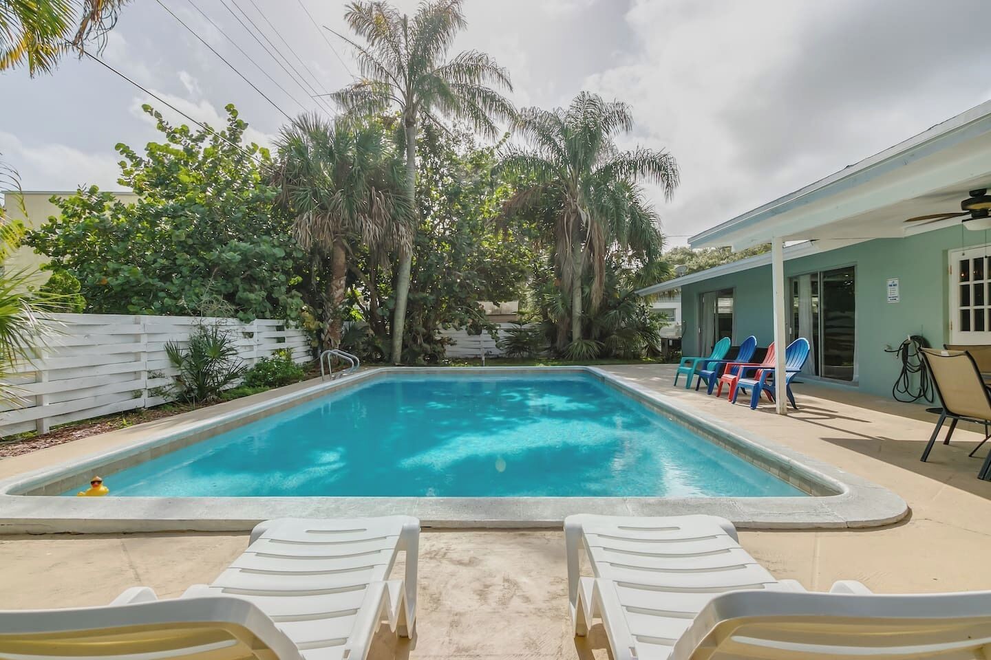 Une piscine rectangulaire dotée d’une terrasse blanche, entourée de palmiers et d’une clôture blanche. On y trouve des chaises longues et un bâtiment à l’extérieur bleu clair.