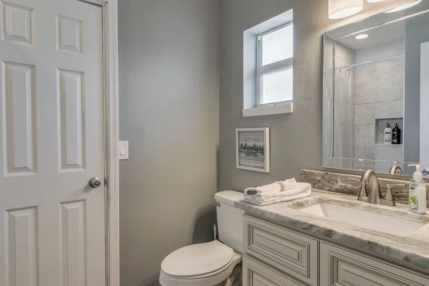A bathroom with a white toilet, a vanity with a marble countertop, a large mirror, and a framed inspirational quote on the wall.