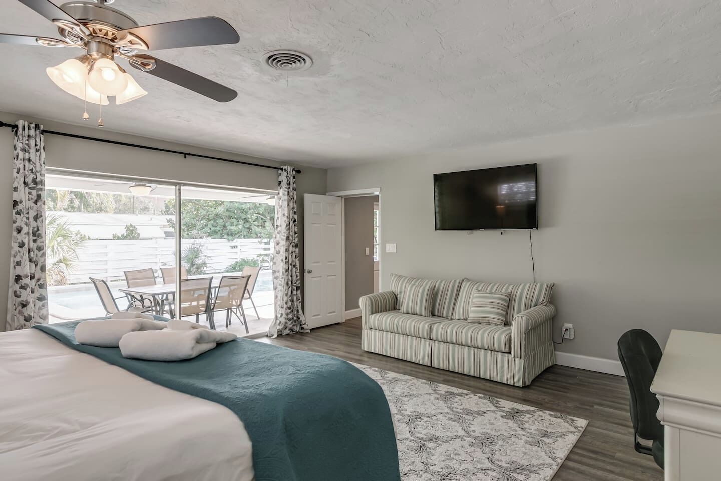 A bedroom with a bed, a ceiling fan, a TV, a sofa, and a view of the outdoors.