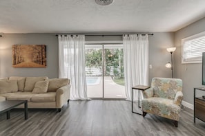 Ein Wohnzimmer mit einem beigefarbenen Sofa, einem gemusterten Sessel, einem Gemälde und Blick durch eine Glastür auf einen Außenbereich.