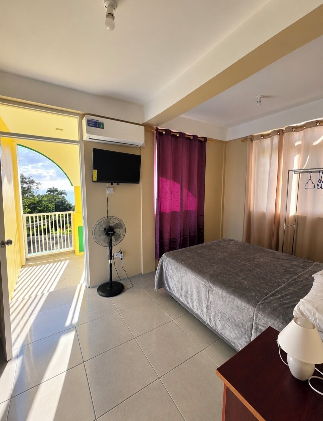 Ein Hotelzimmer mit Bett, Ventilator, Klimaanlage, Fernseher und Blick ins Freie.