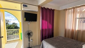 Ein Hotelzimmer mit Bett, Ventilator, Klimaanlage, Fernseher und Blick ins Freie.