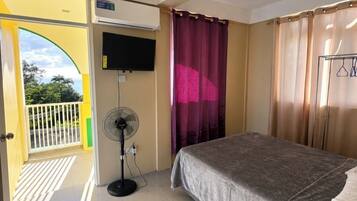 Ein Hotelzimmer mit Bett, Ventilator, Klimaanlage, Fernseher und Blick ins Freie.