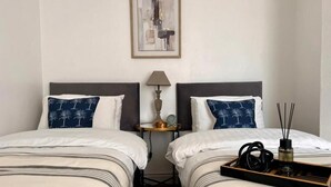 A hotel room with two beds, a nightstand, a lamp, and a tray with a bottle and a remote control. - Spacious Home - Sleeps 5 Persons (Liverpool)