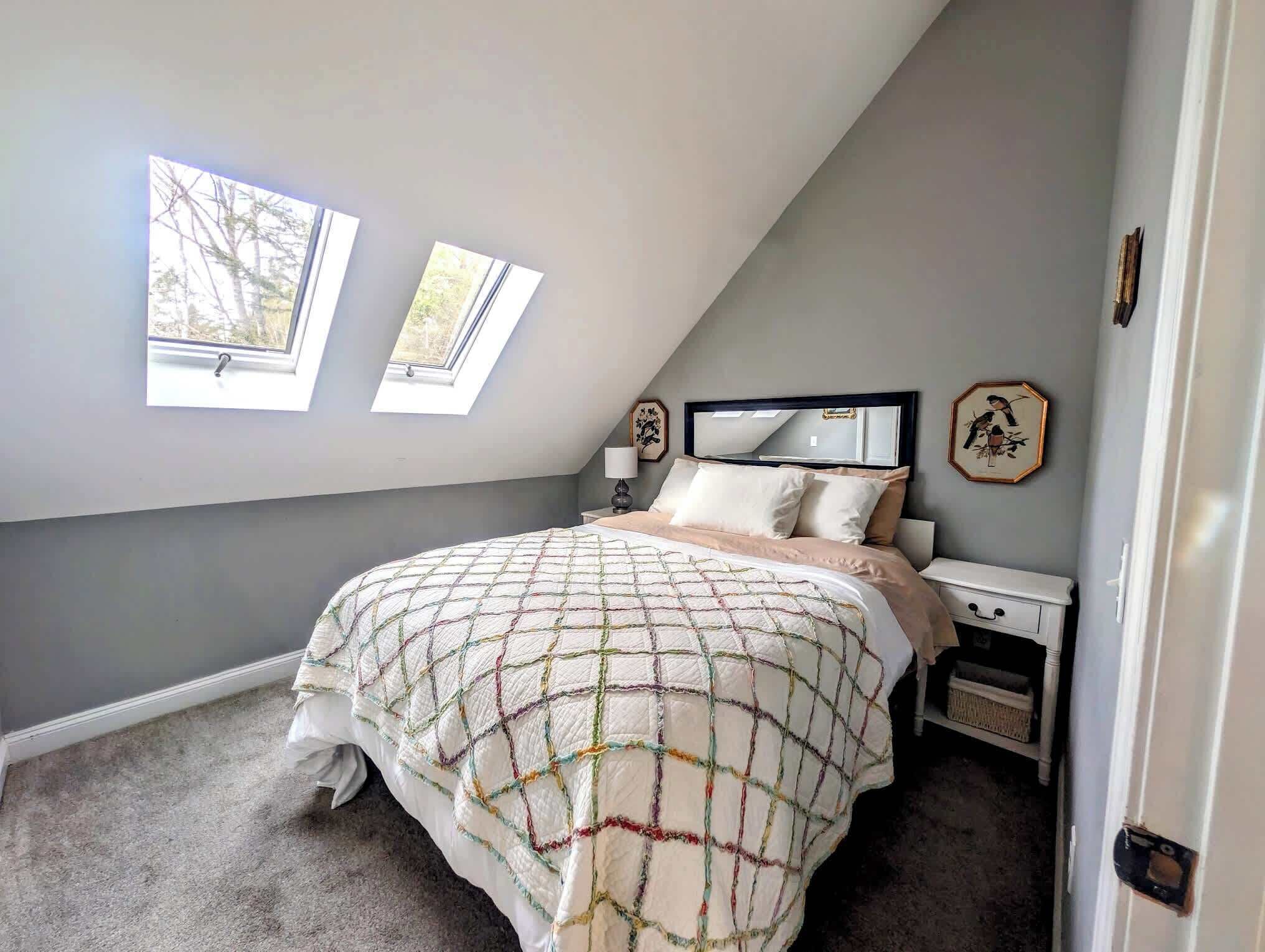 Ein Schlafzimmer mit schrägem Dach, einem Bett mit Bettdecke, zwei Fenstern, einem Nachttisch mit Lampe und einem Spiegel.