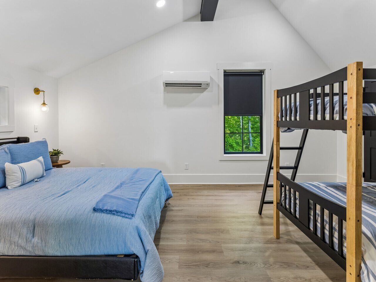 Une chambre avec un lit superposé, un lit avec une literie bleue, une petite fenêtre donnant sur un paysage agréable et un appareil de climatisation.