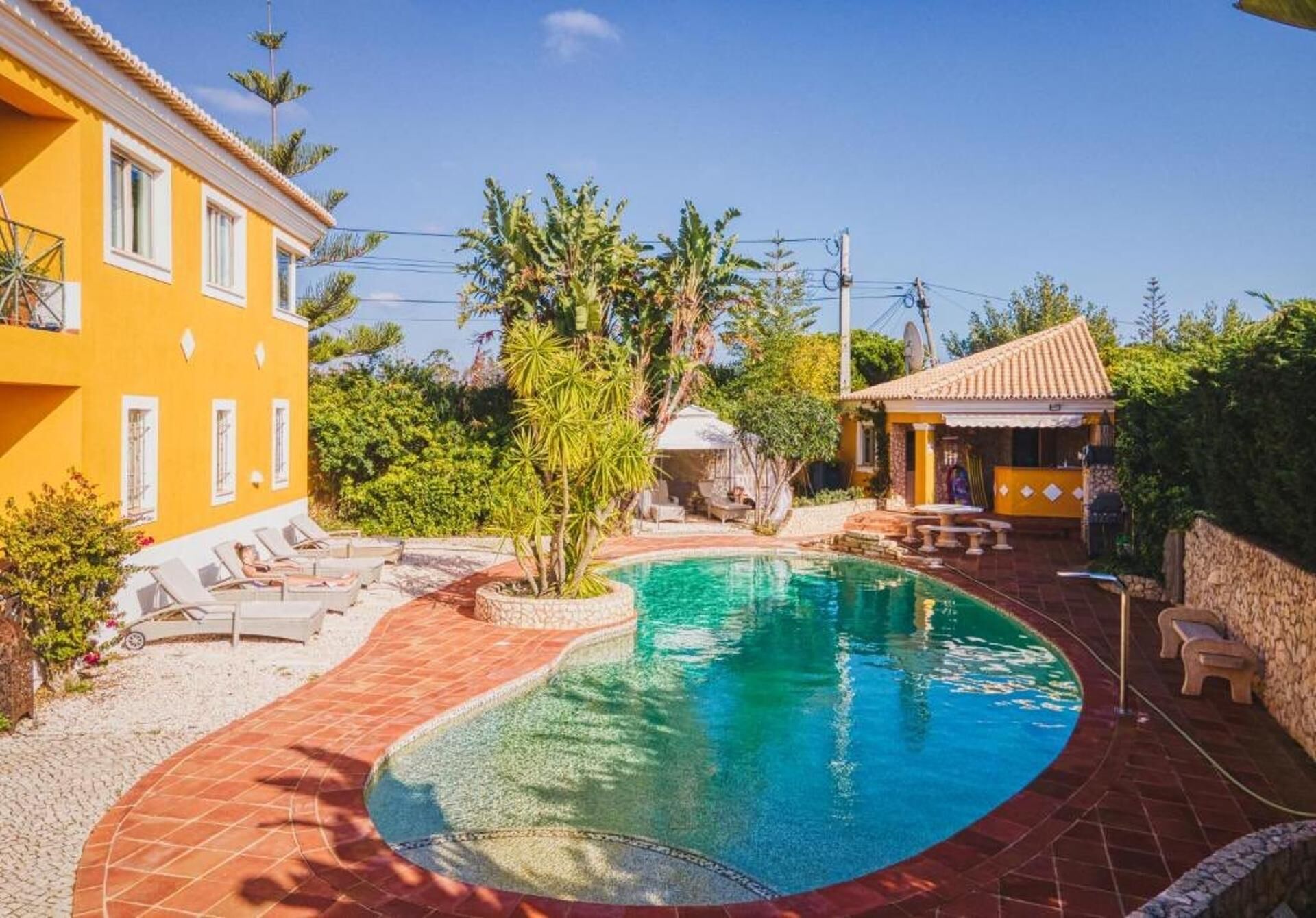 A swimming pool with a tiled deck, surrounded by a yellow building and a smaller structure with a tiled roof.