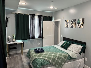 Quarto com cama, escrivaninha, cadeira, janela com cortinas e três obras de arte penduradas na parede.