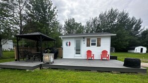 A small white cabin with a porch, two red chairs, a barbecue grill, and a covered patio area. - Cozy cottage River view, close to beaches and amenities. (Grand Barachois)