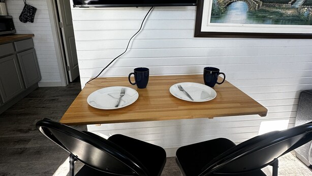 A dining area with a wooden table, two black chairs, a TV mounted on the wall, and a framed landscape painting. - Cozy cottage River view, close to beaches and amenities. (Grand Barachois)