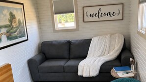 A cozy room with a grey sofa, a teal chair, a white blanket, a framed welcome sign, and a painting of a landscape. - Cozy cottage River view, close to beaches and amenities. (Grand Barachois)