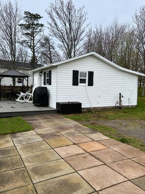 A white single-story house with a covered patio, a black trash bin, and a covered grill area. - Cozy cottage River view, close to beaches and amenities. (Grand Barachois)