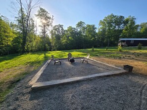 Un espace aménagé avec du gravier, comprenant un foyer, des chaises et une table, entouré d’une terrasse en bois, avec une maison en arrière-plan.