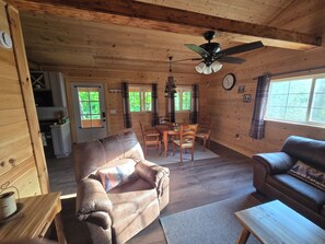 A wooden cabin interior with a dining area, two sofas, a ceiling fan, and a kitchen visible through an open door.