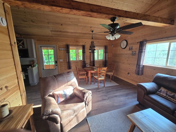 Ein Holzblockhaus-Interieur mit Essbereich, zwei Sofas, Deckenventilator und einer durchgehenden Tür erkennbaren Küche.