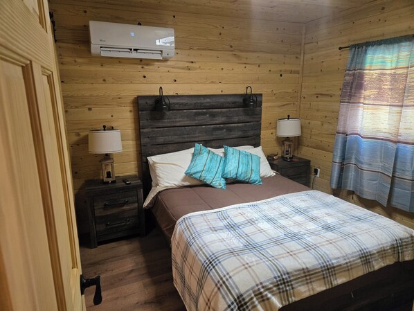 Habitación con paneles de madera, una cama, dos mesitas de noche con lámparas, un equipo de aire acondicionado y una ventana con cortinas.