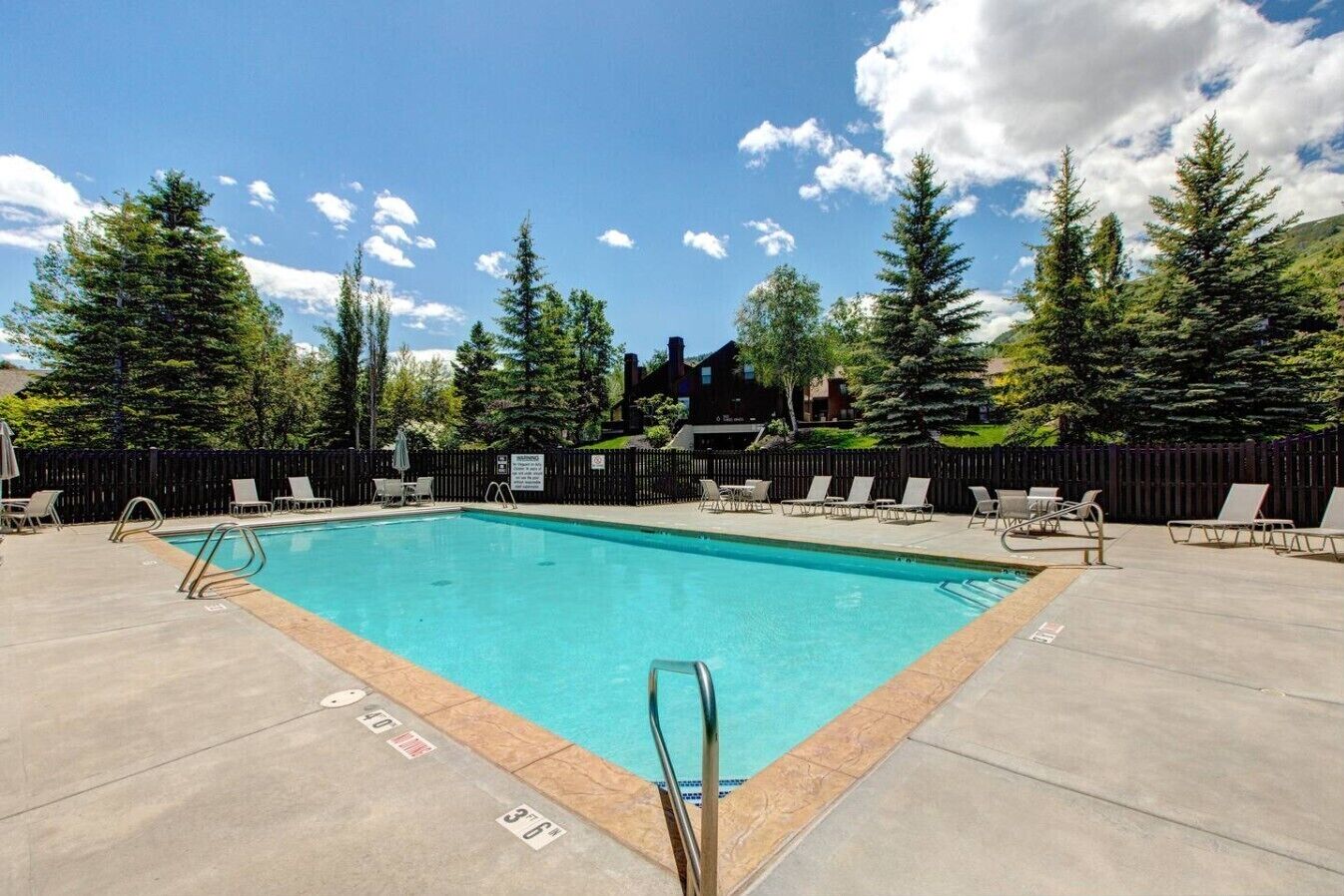 A rectangular swimming pool with a metal ladder, surrounded by lounge chairs and a wooden fence.