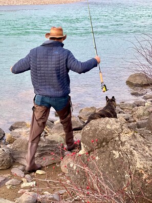 Una persona pescando junto a un cuerpo de agua tranquilo, con rocas y un perro cerca.
