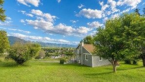 A single-story house with a front porch, surrounded by a well-maintained lawn and trees, set against a backdrop of mountains and a partly cloudy sky.