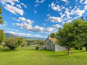A single-story house with a front porch, surrounded by a well-maintained lawn and trees, set against a backdrop of mountains and a partly cloudy sky.
