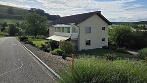 Ein Haus mit Wintergarten, umgeben von Grünpflanzen und einer Straße.