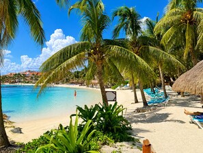 A beach with palm trees, a clear blue sea, and a thatched-roof structure. - Marina view 3BR pool 7min walk to beach (Puerto Aventuras)