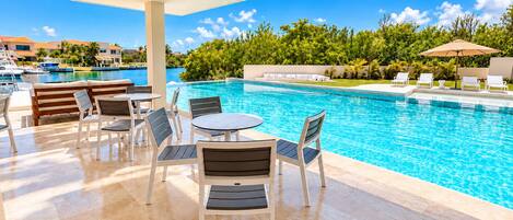 An outdoor pool area with lounge chairs and tables, a clear view of the pool, and a backdrop of greenery and buildings.