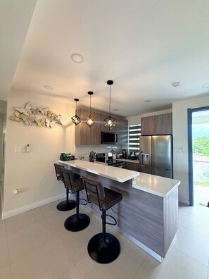 Een moderne keuken met een centraal eiland, barkrukken, hanglampen en uitzicht op de omgeving.