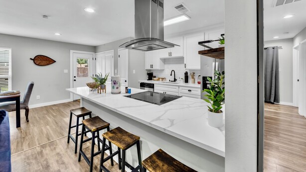 A modern kitchen with white cabinetry, a central island with stools, a stainless steel range hood, and a dining area with a blue sofa and wooden chairs. - 3BR Home Near Outlets & Event Center ,
Pet Friendly, Backyard (Allen)
