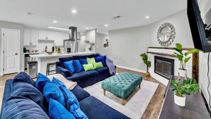 A modern living room with a dark blue sofa, a teal ottoman, and a flat-screen TV. - 3BR Home Near Outlets & Event Center ,
Pet Friendly, Backyard (Allen)