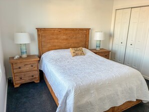 Ein ordentlich bezogenes Bett mit weißer Decke, zwei Nachttische mit Lampen, ein Holz-Kopfteil, eine kleine Schublade und ein Kleiderschrank mit Türen.