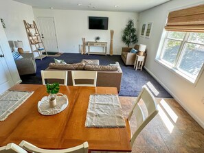 Ein Essbereich mit Holztisch, weißen Stühlen und einer zentralen Pflanze. Im Hintergrund ist ein Wohnzimmer mit Sofa und Fernseher zu sehen.