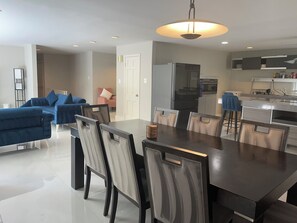 Ein modernes Wohnzimmer mit einem dunkelholzigen Esstisch, hellen Stühlen, einem blauen Sofa und einer Küchenzeile mit Kühlschrank und Herd.